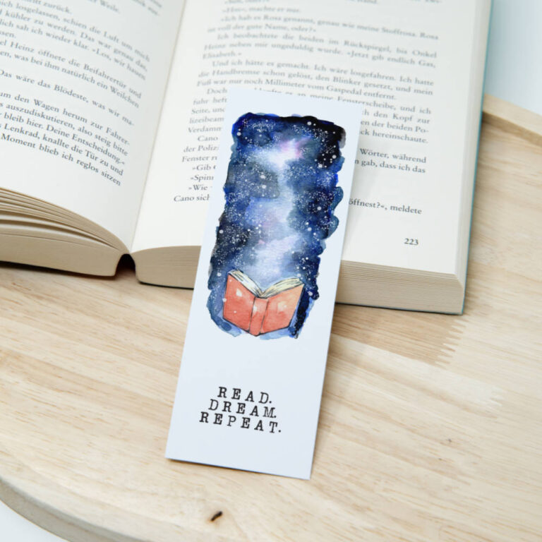 Aquarell-Lesezeichen mit nächtlichem Himmel in Blau- und Lilatönen, aufgeschlagenem Buch und dem Spruch Read Dream Repeat auf Papier.