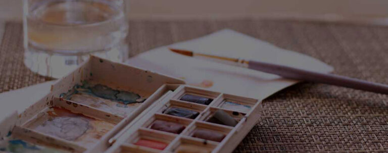 Aquarellfarben, Pinsel und Wasserglas auf Rattantisch