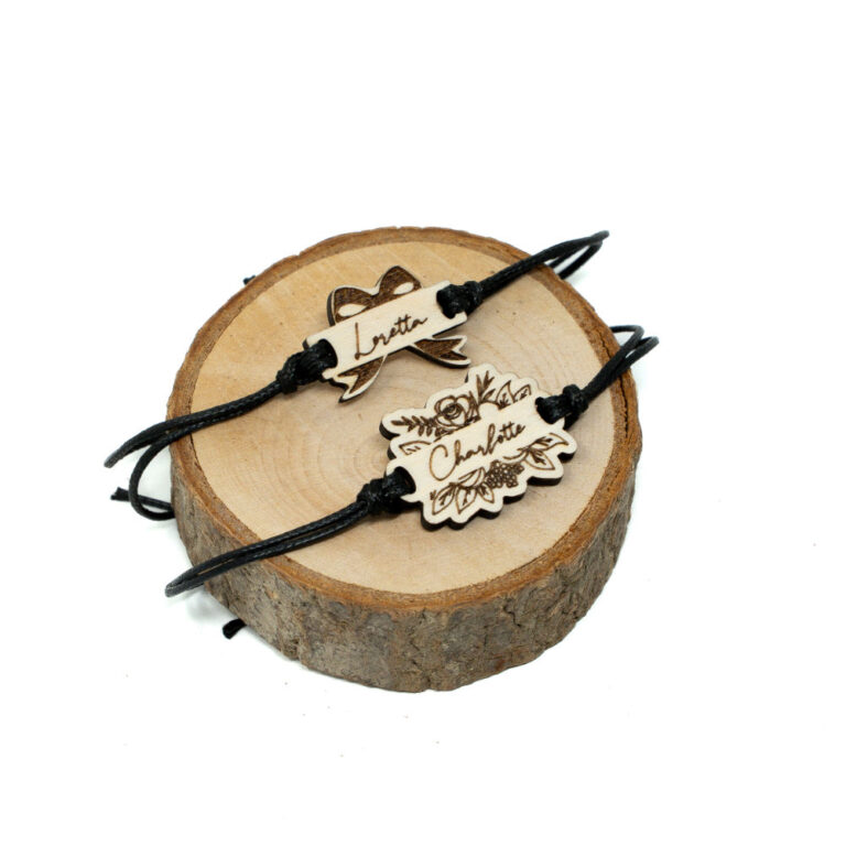 Zwei verstellbare schwarze Kordel-Armbänder auf Holzscheibe, oben Schleifenarmband mit Namen Loretta, unten florales Brautarmband mit Namen Charlotte, beide aus naturbelassenem Holz mit Lasergravur.