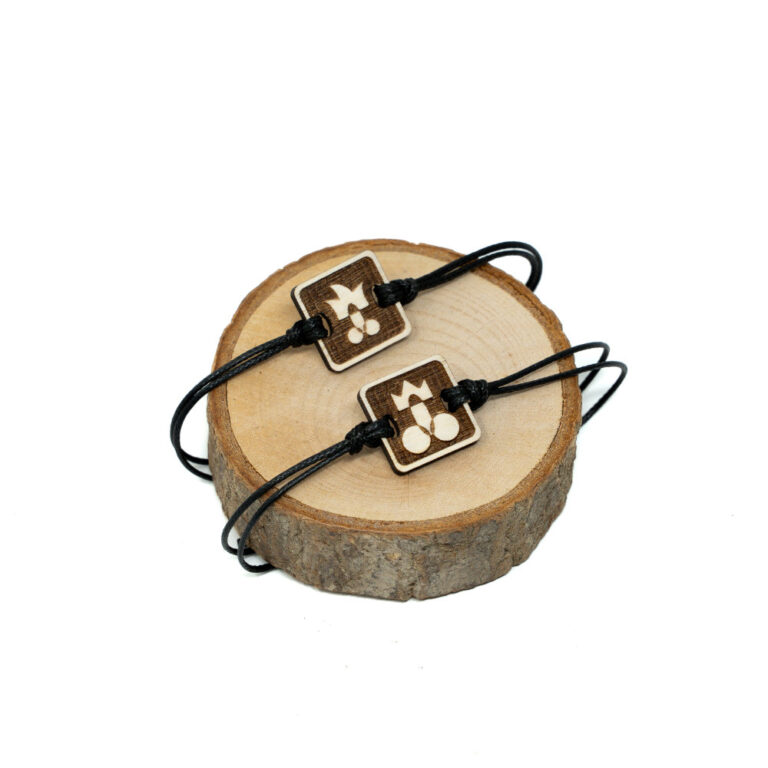 Zwei verstellbare schwarze Kordel-Armbänder auf Holzscheibe mit lasergeschnittenen quadratischen Holzanhängern, oben Pimmel mit Krone für den Bräutigam, unten Pimmel mit Narrenmütze für das Team.