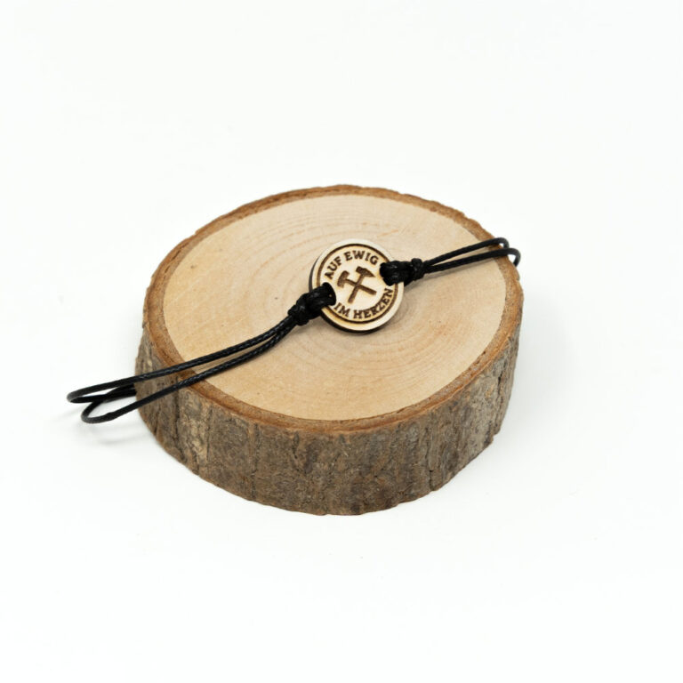 Verstellbares schwarzes Kordel-Armband mit rundem Holzanhänger und graviertem Hacke-Schlegel-Symbol mit Schriftzug Auf ewig im Herzen auf hellem naturbelassenem Holzhintergrund.