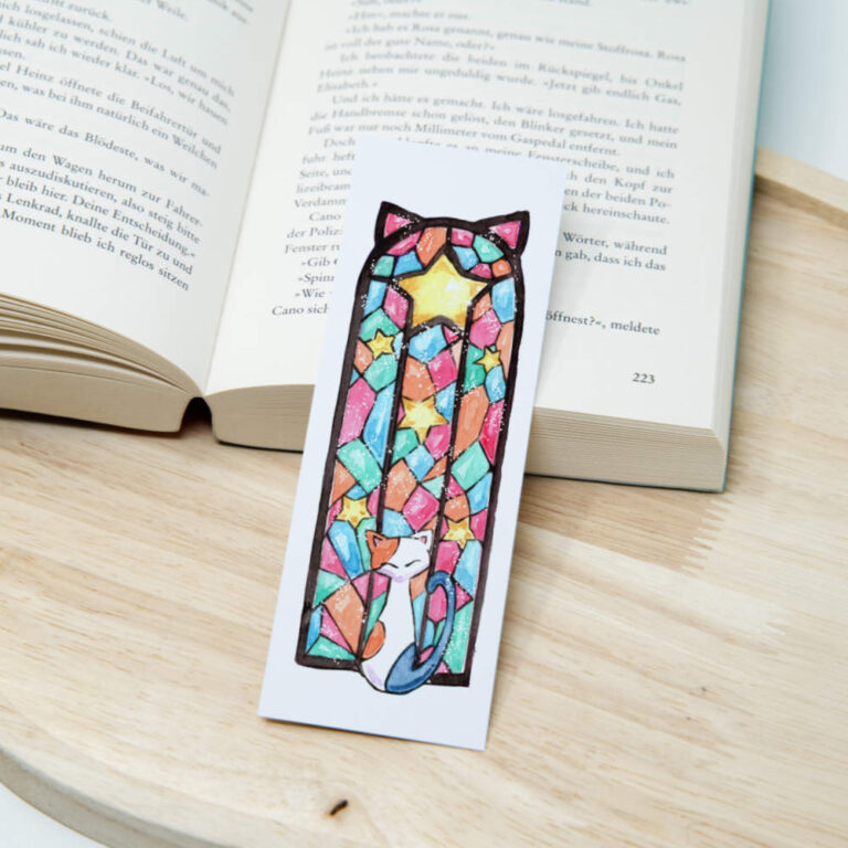 Aquarell-Lesezeichen mit einer Katze die vor einem bunten kirchenähnlichen Fenster in leuchtenden Farben sitzt, stimmungsvolles weihnachtliches Motiv auf Papier.
